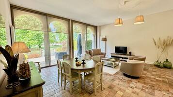 A spacious living area with a dining table, chairs, a sofa, and a TV. Large windows provide a view of the outdoors.