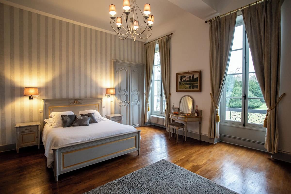 Een slaapkamer met een groot bed, nachtlampjes, een dressoir, een bureau en een raam met gordijnen.