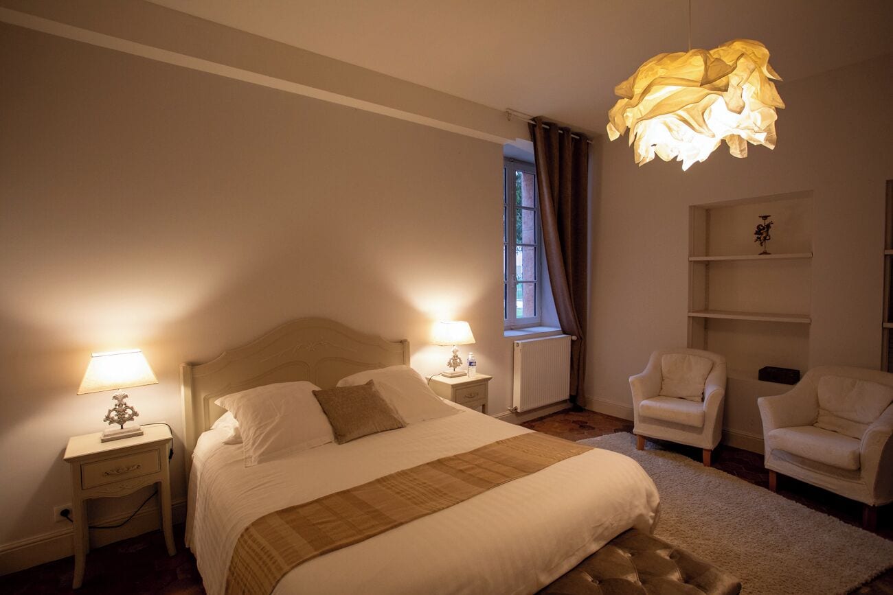 A bedroom with a bed, bedside lamps, a window with curtains, and two armchairs.
