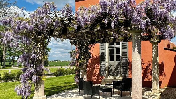 A red building with a tiled roof, a pergola covered in purple flowers, and a patio with outdoor furniture.