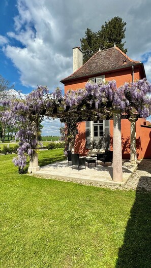 A red building with a tiled roof, a pergola covered in purple flowers, and a patio with outdoor furniture. - Château de Quincey (Quincey)