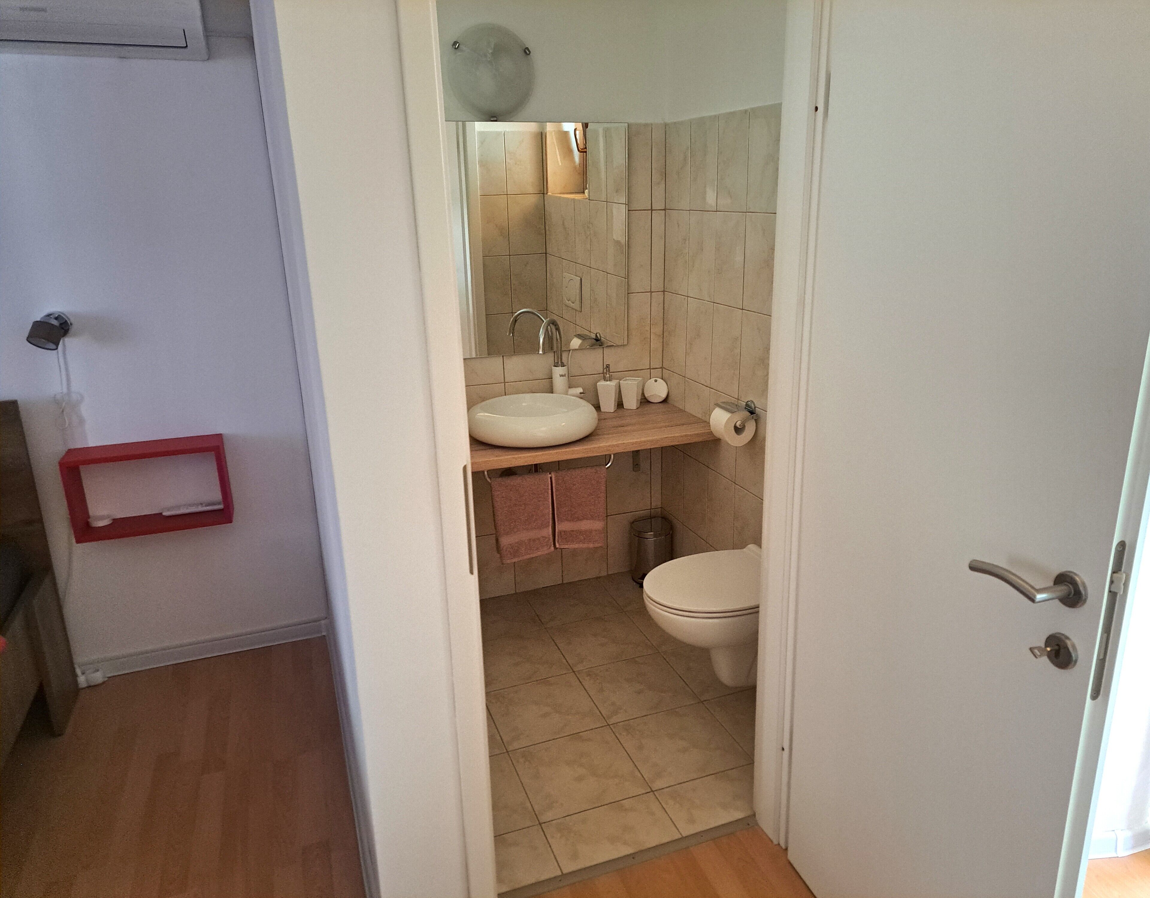 Ein Badezimmer mit einem weißen Waschbecken, einer Toilette und einem Spiegel über dem Waschbecken. An der Wand befindet sich ein roter Handtuchhalter und ein Mülleimer unter dem Waschbecken.