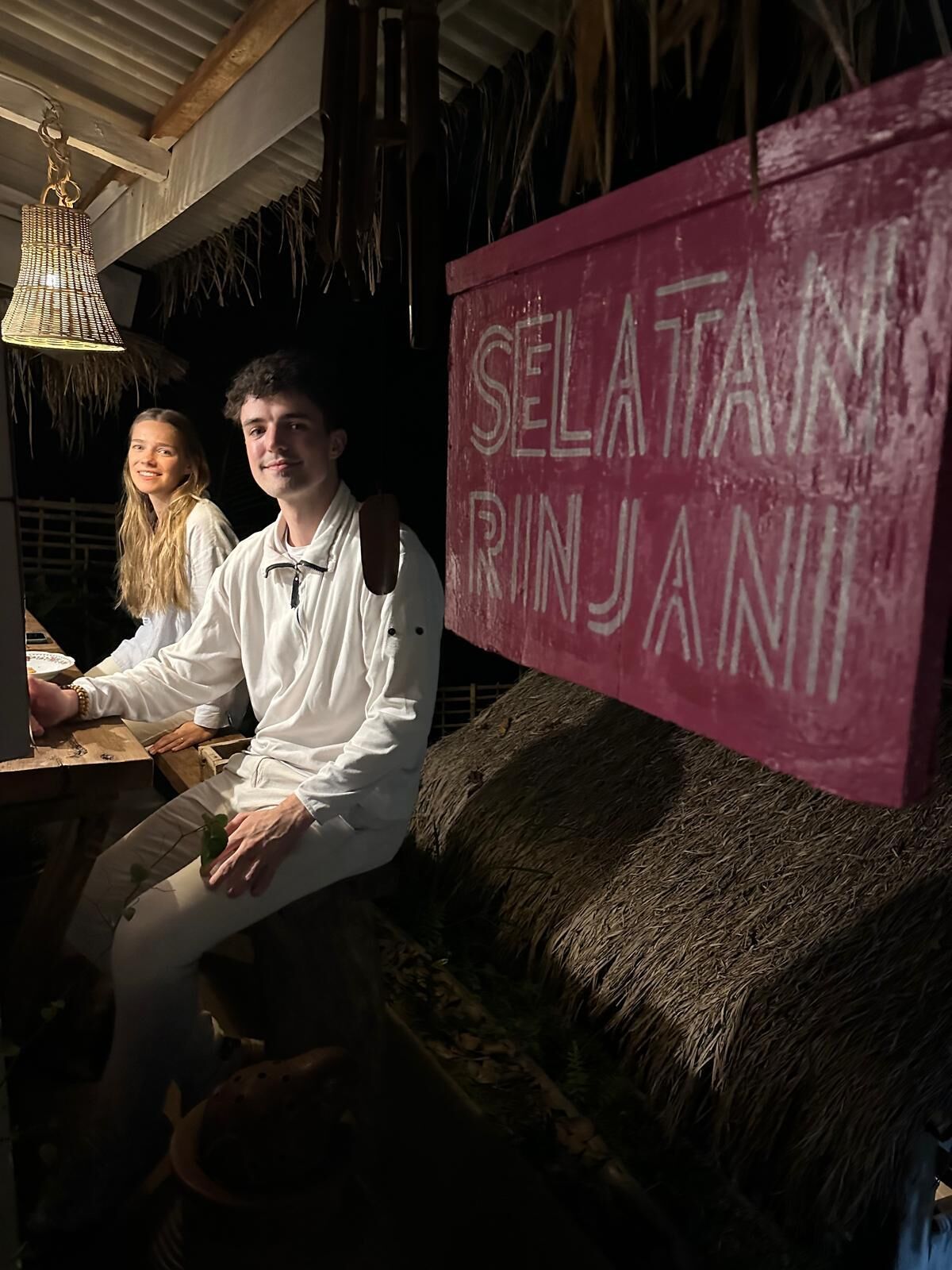 A rustic interior with a thatched ceiling, a hanging light fixture, and a sign reading "SELATAN RINJANI".