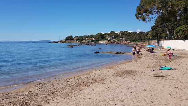 Una playa con gente tomando el sol y nadando, un velero en el agua y casas al fondo.