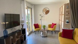 Una sala de estar con televisor, sofá amarillo, cojines rojos, una mesita con una planta y un reloj en la pared.
