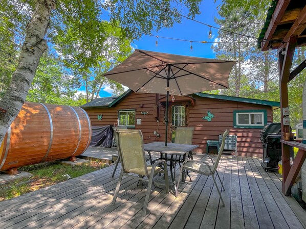 A wooden deck with a table and chairs, a large barrel, and a small cabin with a green roof.