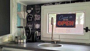 Een keuken met een spoelbak, een koffiezetapparaat en een broodrooster. Er is een raam met een bord waarop staat: Come in we're OPEN.