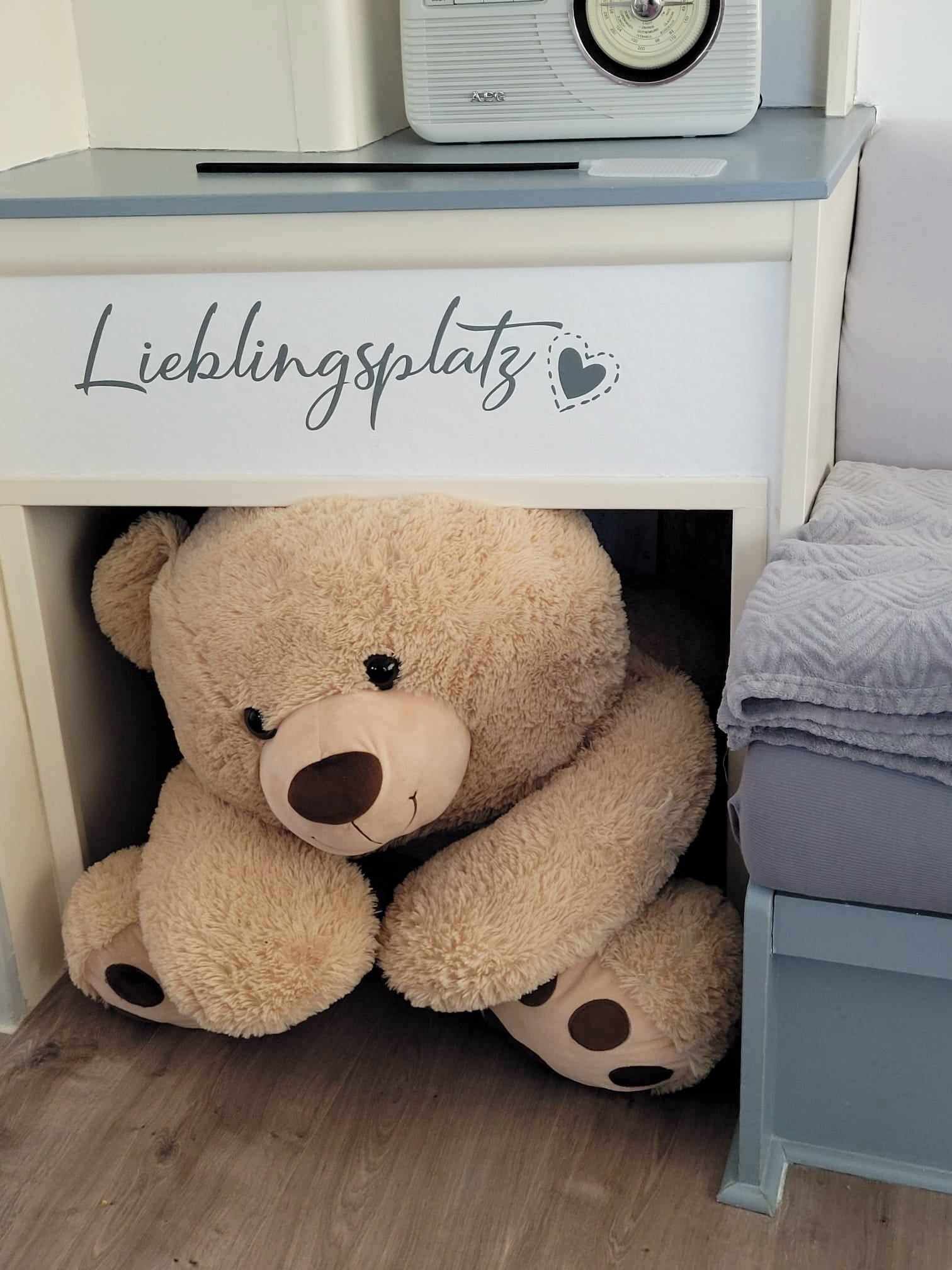A bed with a white headboard, a bedside table with a radio, and a teddy bear under the bed.