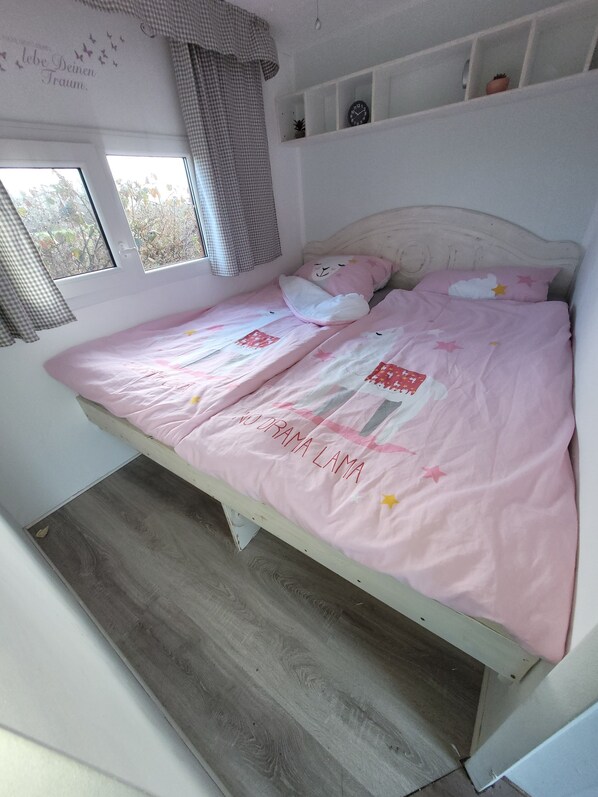 A bed with pink bedding and a quilt with 'NO DRAMA LAMA' text, a window with a view of trees, and a shelf with a clock and a plant.