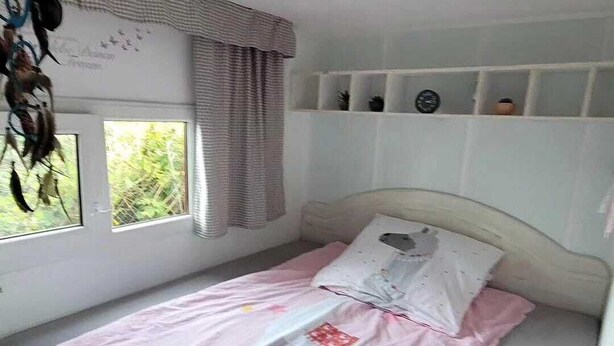 Un llit individual amb llençols rosats i una almoina blanca, una finestra amb cortines, i una estant amb un rellotge i objectes decoratius.