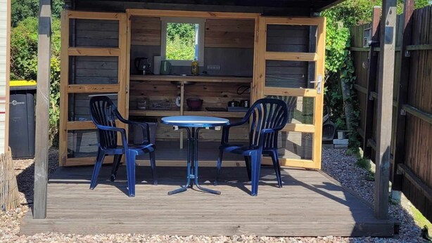 Een houten buitenkeuken met een tafel en stoelen, omgeven door een houten terras en een pergola met verlichting.