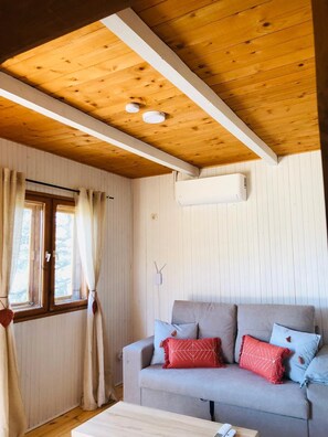 A cozy room with a wooden ceiling, a grey sofa with patterned cushions, a window with curtains, and an air conditioning unit.