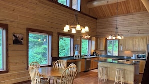 A wooden cabin interior with a dining table, chairs, a sofa, a kitchen with a bar, and large windows.