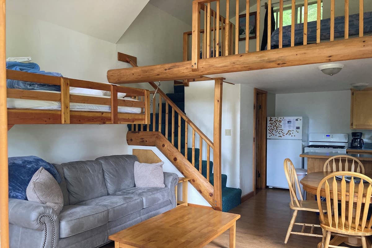 A cozy room with a bunk bed, a gray sofa, a wooden table, and a kitchen area with a stove and chairs.