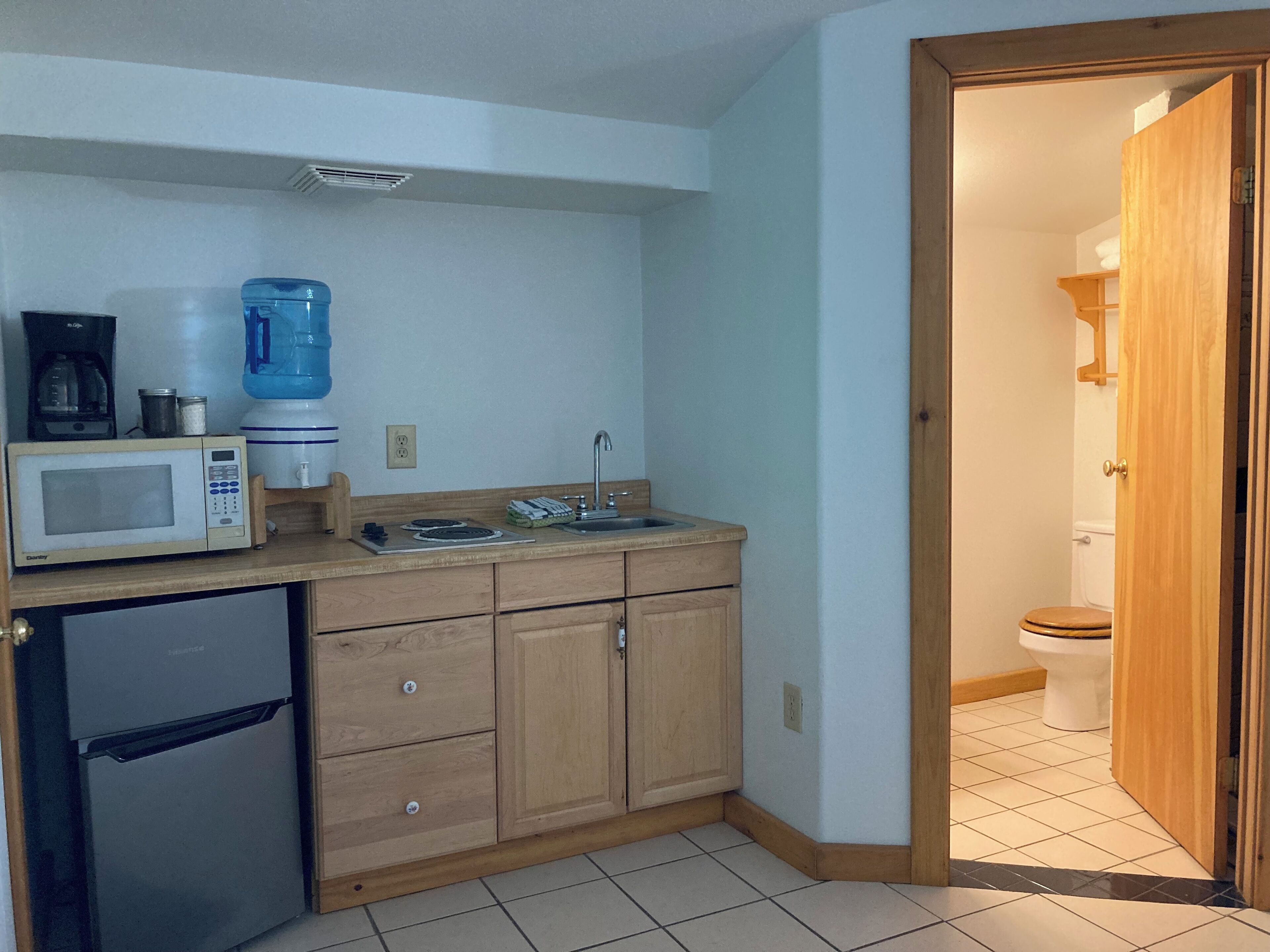 A compact kitchen with a microwave, coffee maker, and water dispenser. A small refrigerator is on the left, and a wooden cabinet with a sink is on the right. A wooden door leads to a bathroom with a toilet.