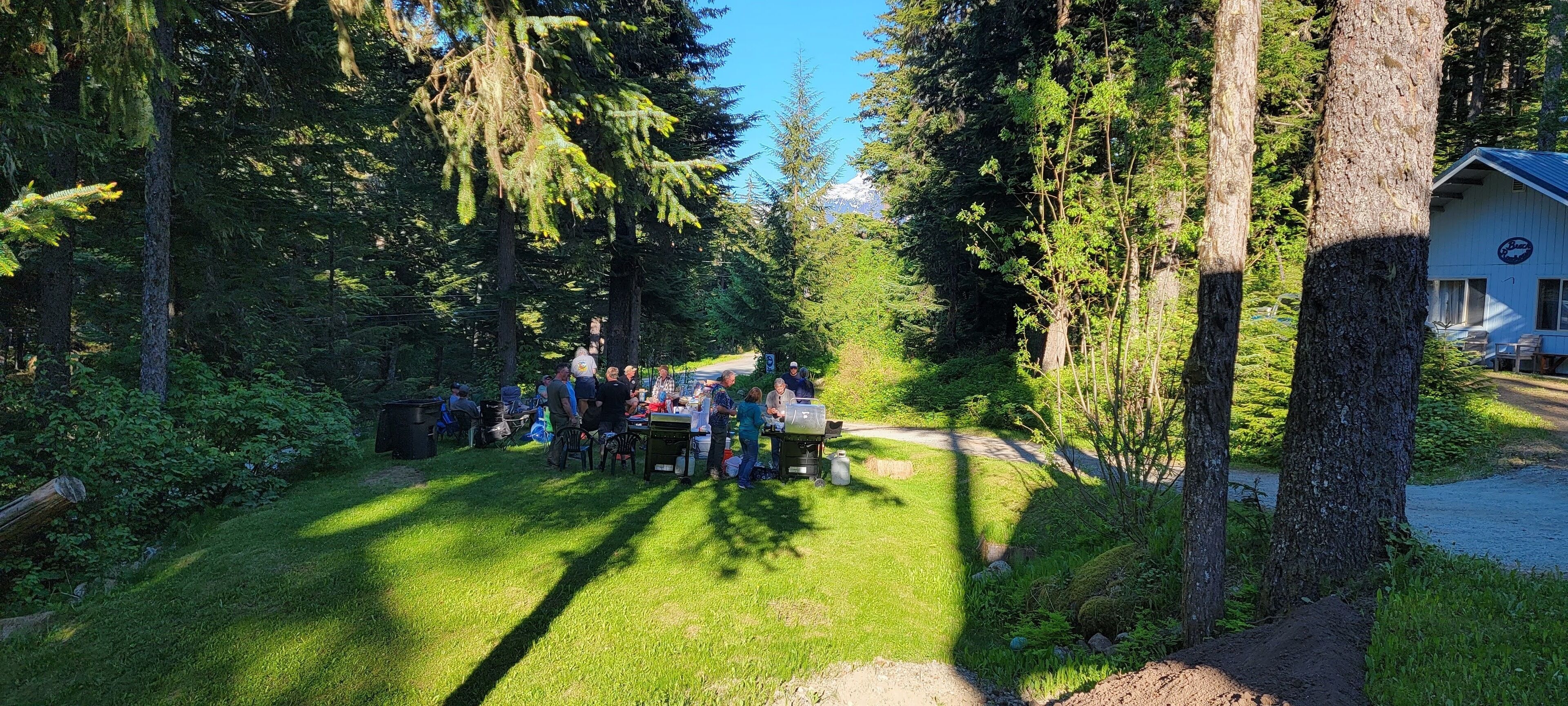 A group of people gathered around a table in a forested area with a cabin in the background.