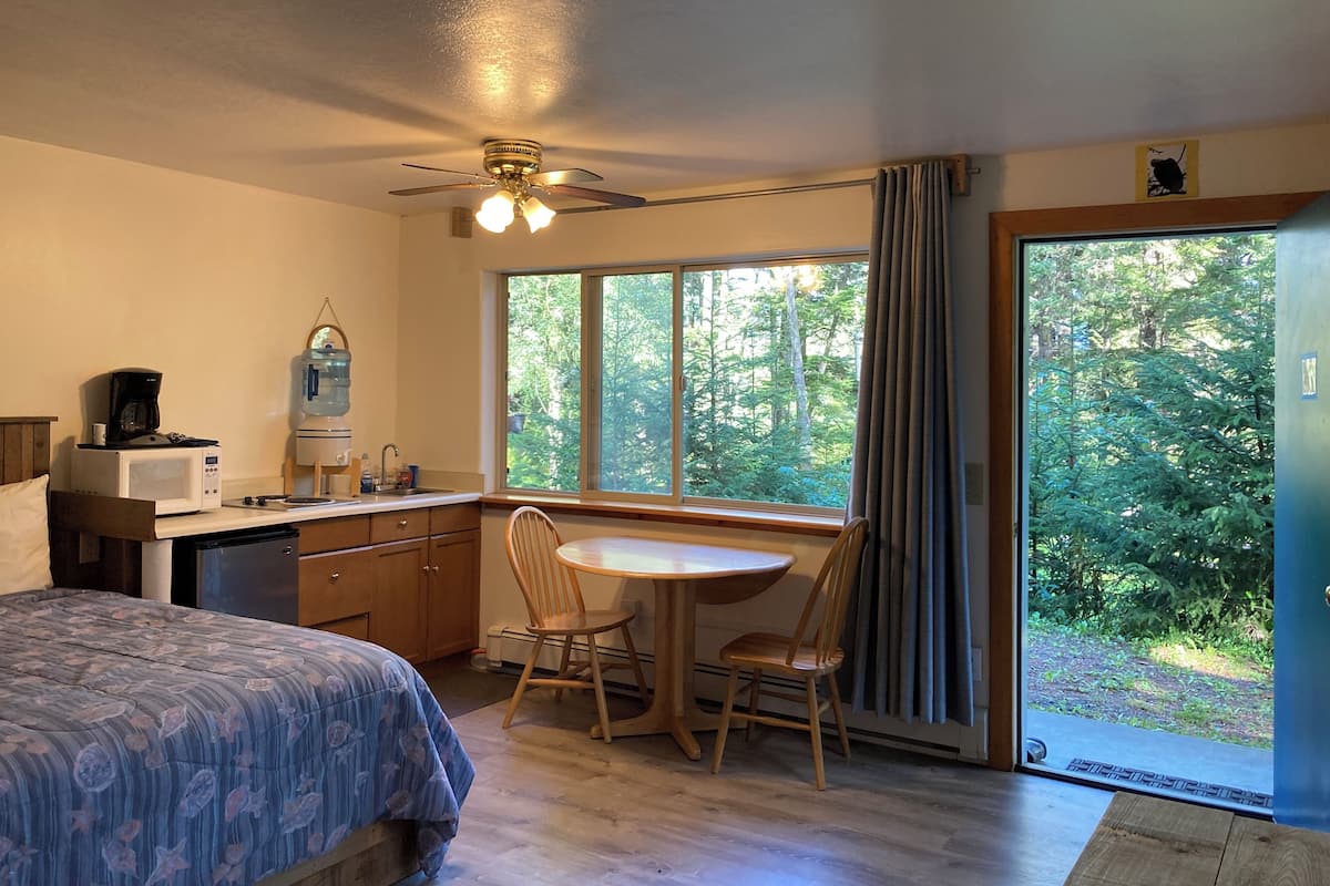 A compact room with a bed, a small kitchenette, a dining table with chairs, and a large window offering a view of a forest.