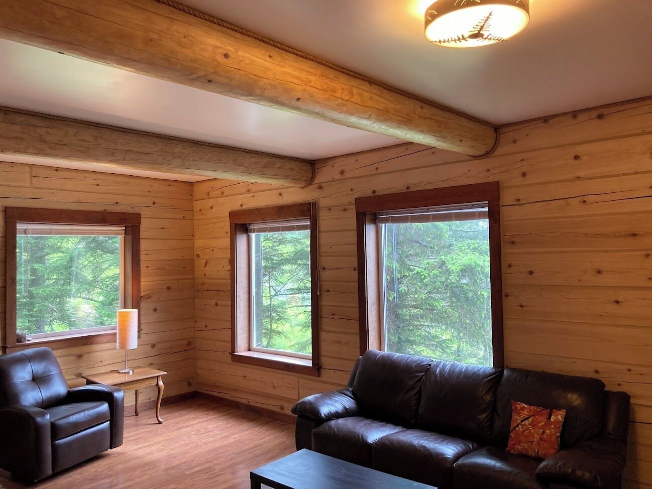 A log cabin interior with a leather sofa, a small table, and large windows offering a forest view.