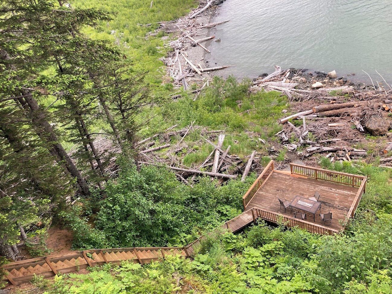 A wooden deck with a table and chairs, surrounded by dense greenery and a body of water.