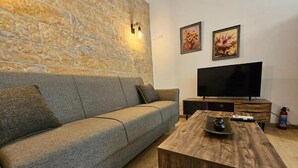 A living room with a stone wall, a grey sofa, a wooden coffee table, a flat-screen TV, and two framed pictures. - Honeywell Comfort Stay (Markopoulo Mesogaias)