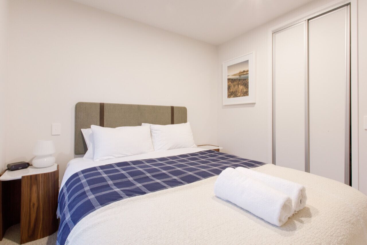 Een net opgemaakt bed met een blauw-wit geruite deken, een wit kussen en een opgerolde witte handdoek. Er staat een houten nachtkastje met een witte lamp en een klein elektronisch apparaat. Daarboven hangt een ingelijste foto aan de muur.