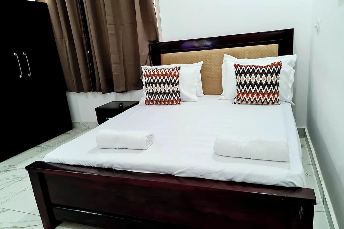 A neatly made bed with white linens and two patterned pillows, a dark wooden headboard, and a marble-tiled floor.
