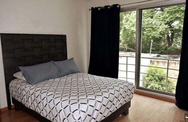 A bedroom with a patterned bedspread, a dark headboard, and a large window with curtains.