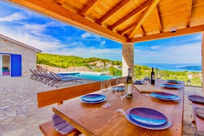 An outdoor dining area with a wooden table set for a meal, overlooking a pool and a scenic view of hills and the sea.