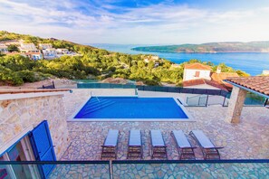 A swimming pool with lounge chairs, a stone patio, and a view of the sea and hills.