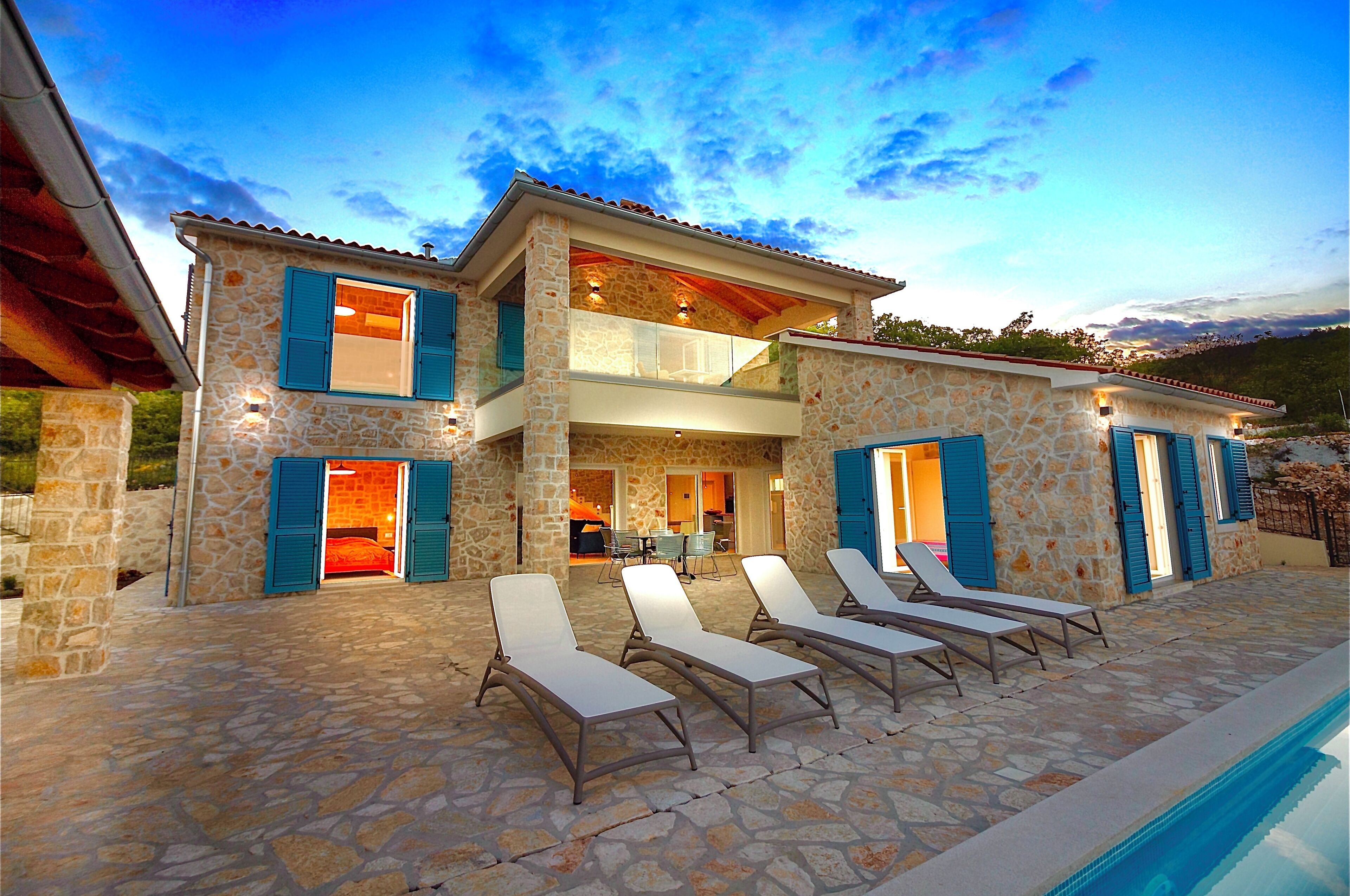 A two-story stone house with a pool, outdoor seating, and a view of the surrounding landscape.