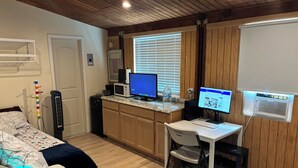 Une pièce compacte comprenant un coin cuisine, un lit, un bureau avec une chaise et une fenêtre avec des stores.