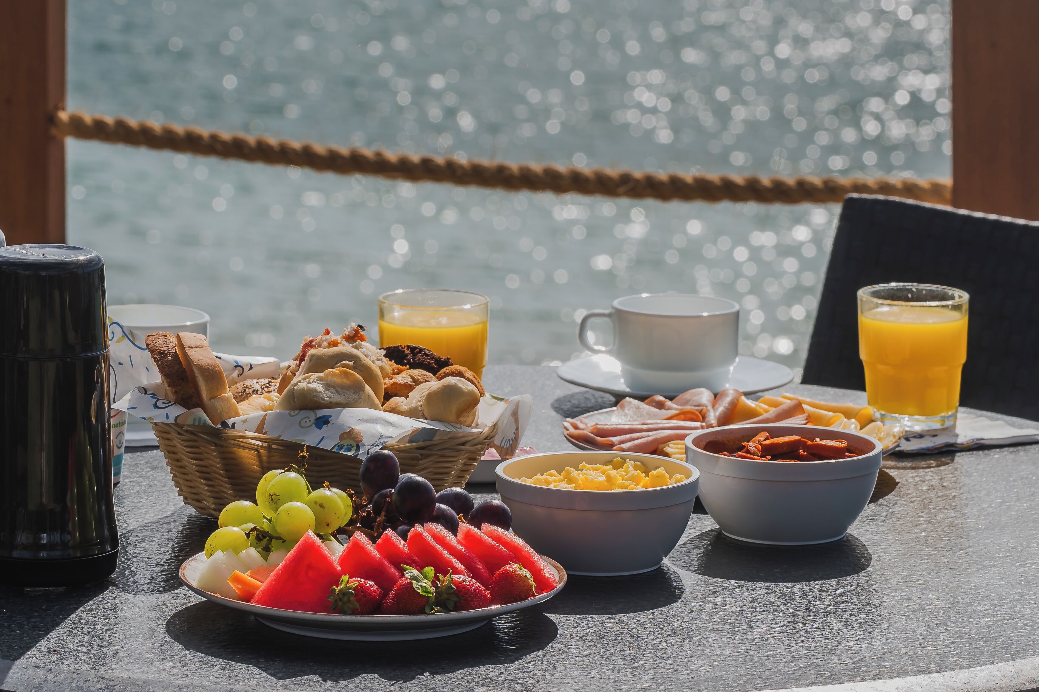 Una mesa de desayuno con diferentes alimentos y bebidas, una cesta de pan y vista al agua.