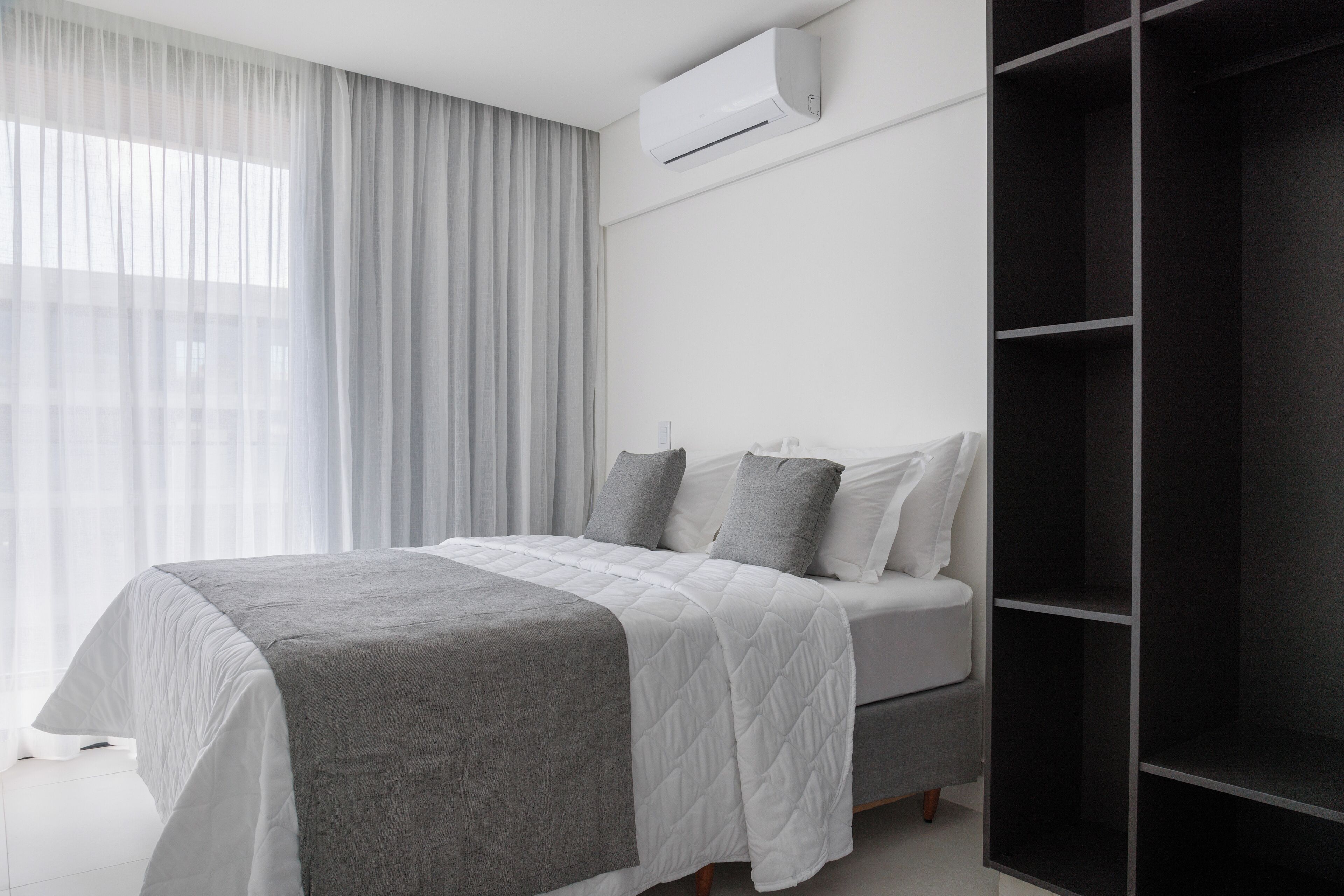 A modern bedroom with a bed, grey and white bedding, a grey throw blanket, a white air conditioner, and a large wardrobe.