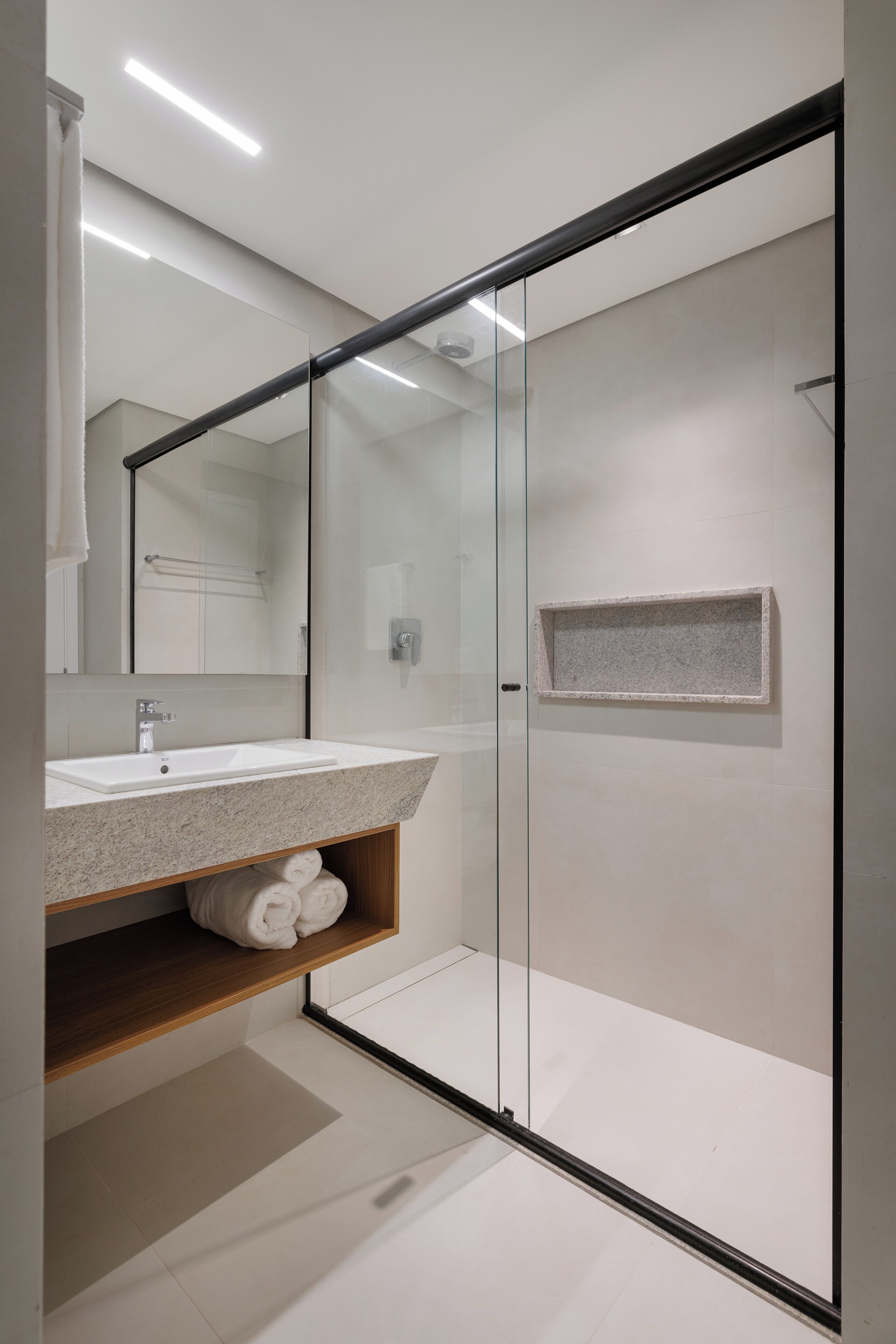 Un baño moderno con cabina de ducha de vidrio, un lavabo blanco con repisa de madera y un espejo grande.