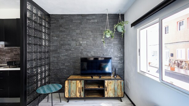 A modern living room with a TV on a wooden cabinet, a blue sofa, and a large window with a view of buildings. - Estudio en La Jaca, a 3 Minutos de la Playa, en un Lugar Ideal Para Desconectar (La Jaca)