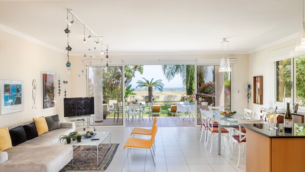 A modern living room with a sofa, TV, and dining area with chairs and a table. - Larnaca Chelidonia Seafront Villa (Meneou)