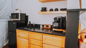 Una cocina compacta con gabinetes de madera, un refrigerador negro, un microondas, una cafetera y una repisa con tazas y platos.