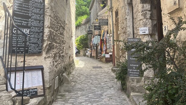 Une ruelle étroite pavée de pierres, bordée de bâtiments en pierre, avec des enseignes indiquant les commerces locaux, et un ciel d’un bleu limpide.