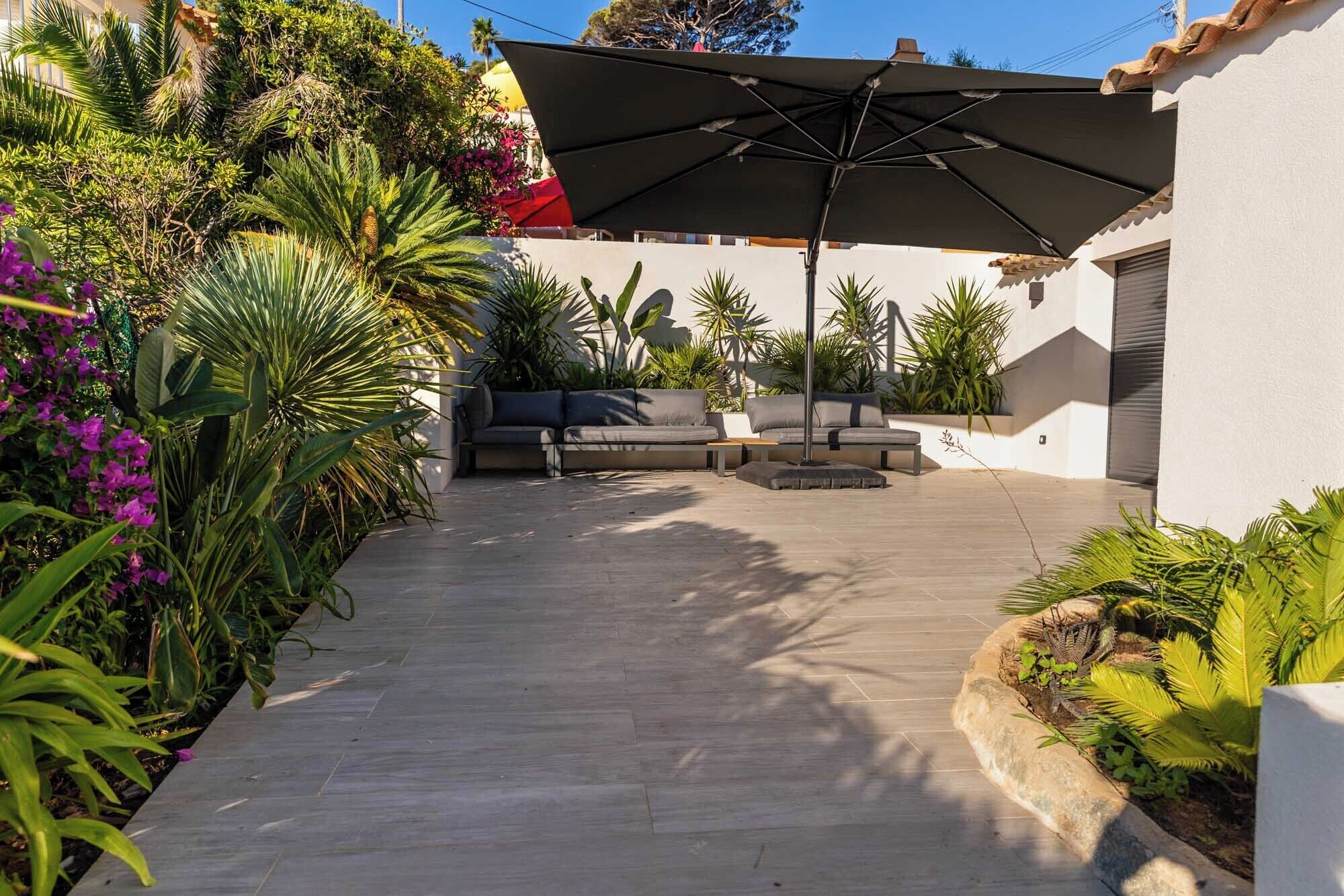 Eine Terrasse mit einer Couch und einem Sonnenschirm, umgeben von üppigem Grün und Pflanzen.