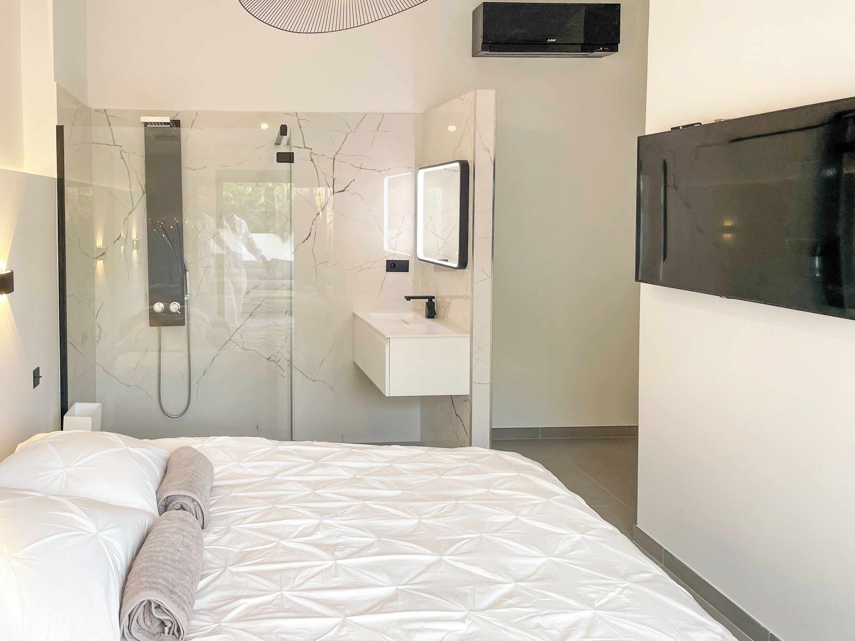 A modern bedroom with a large bed, a built-in shower, a white vanity, and a flat-screen TV mounted on the wall.