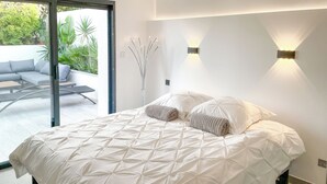 Une chambre à coucher moderne avec un grand lit, une porte vitrée donnant sur un patio extérieur et un luminaire mural minimaliste.