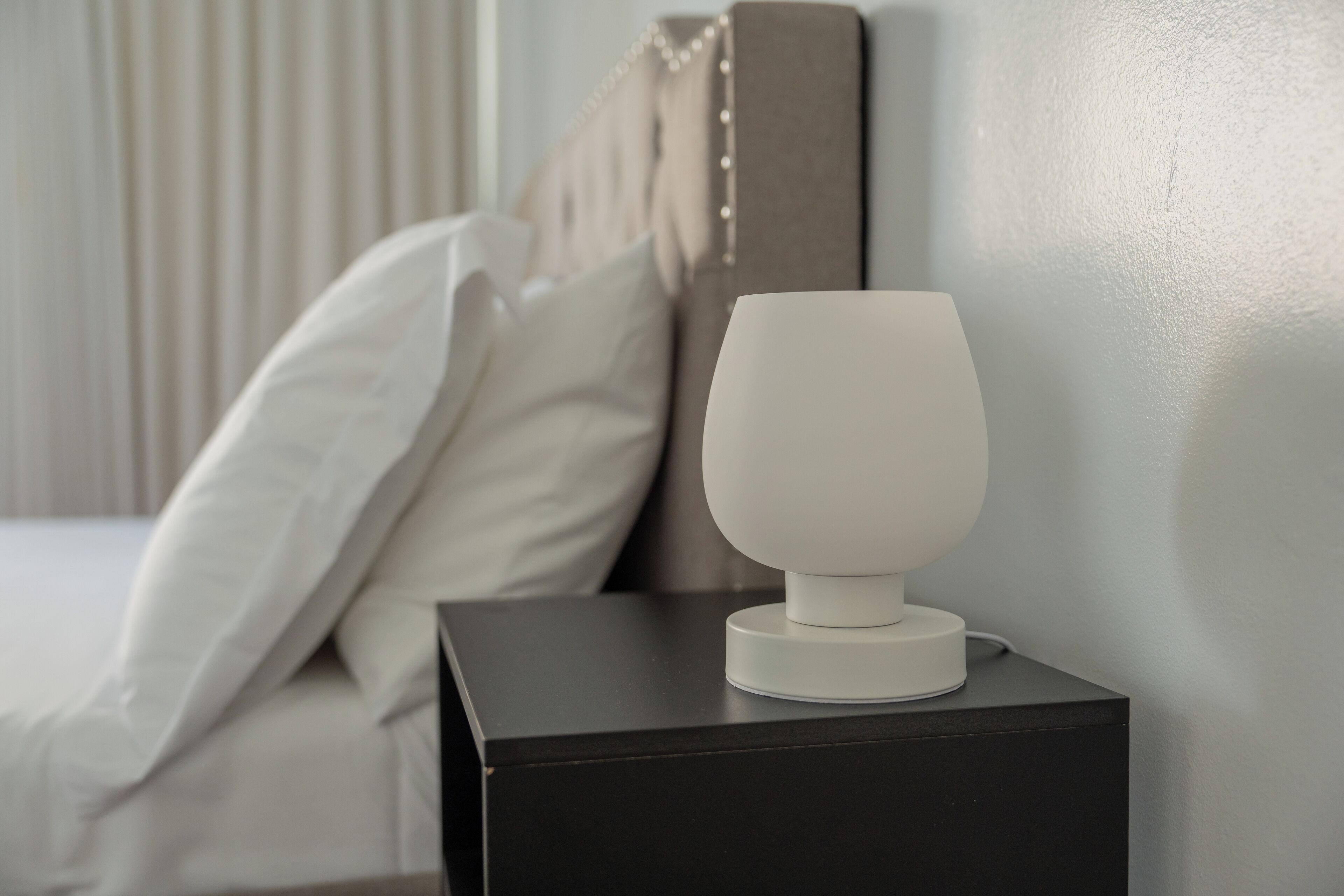 A bedside table with a white lamp, a bed with white bedding, and a headboard with vertical blinds.