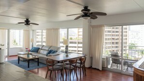 Ein geräumiges Wohnzimmer mit großem Fenster, Deckenventilator, Esstisch mit Stühlen und einer Couch mit Kissen.