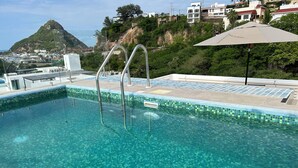 A clear blue pool with metal ladders, a tiled edge, and a view of a mountain and buildings in the background. - Carrizo apartment (Mazatlán)