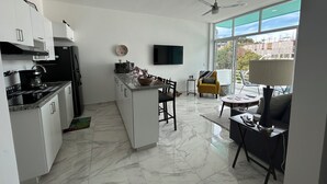 A modern kitchen with white cabinets, a black refrigerator, and a coffee maker. There is a dining area with a yellow armchair and a table. A ceiling fan is visible, and a TV is mounted on the wall. - Carrizo apartment (Mazatlán)