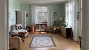 A cozy room with a wooden floor, a large window with sheer curtains, wicker furniture, a bookshelf, and a small table with a lamp. - Summer House in Swedish Archipelago (Stockholms län)