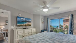 Ein Schlafzimmer mit Bett, Deckenventilator, Fernseher auf einer Kommode, einem Strandbild und einem Balkon mit Blick auf Gebäude und Grünflächen.