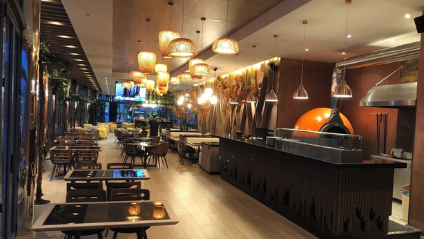 A modern restaurant interior with multiple dining tables and chairs, hanging pendant lights, and a central counter with a pizza oven. - Riverside rooms at Hotel Fieri (Fier)
