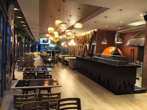 Interior moderno de restaurante com várias mesas e cadeiras, luminárias pendentes e um balcão central com forno a lenha para pizza.