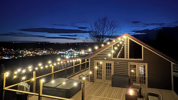 Una terraza de madera con luces colgantes, un espacio cubierto para sentarse y vistas a un pueblo al anochecer.